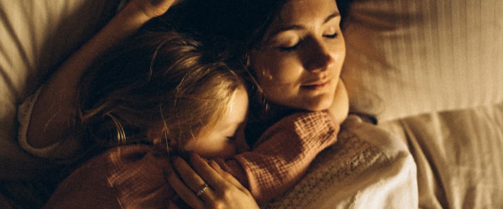 Mother and daughter cuddling happily on a comfortable white bed with a yellow hue from the electric fire