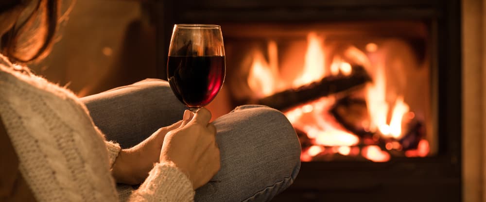 Woman wearing comfortable woolen clothes holding a full glass of red wine while curled up in front of a warm wood fire