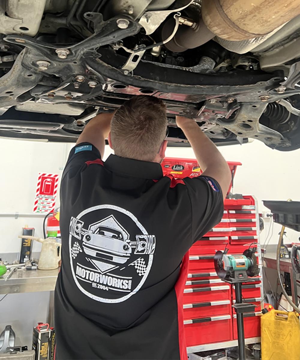  Expert mechanic in a Motor Works Group branded shirt servicing  an elevated car