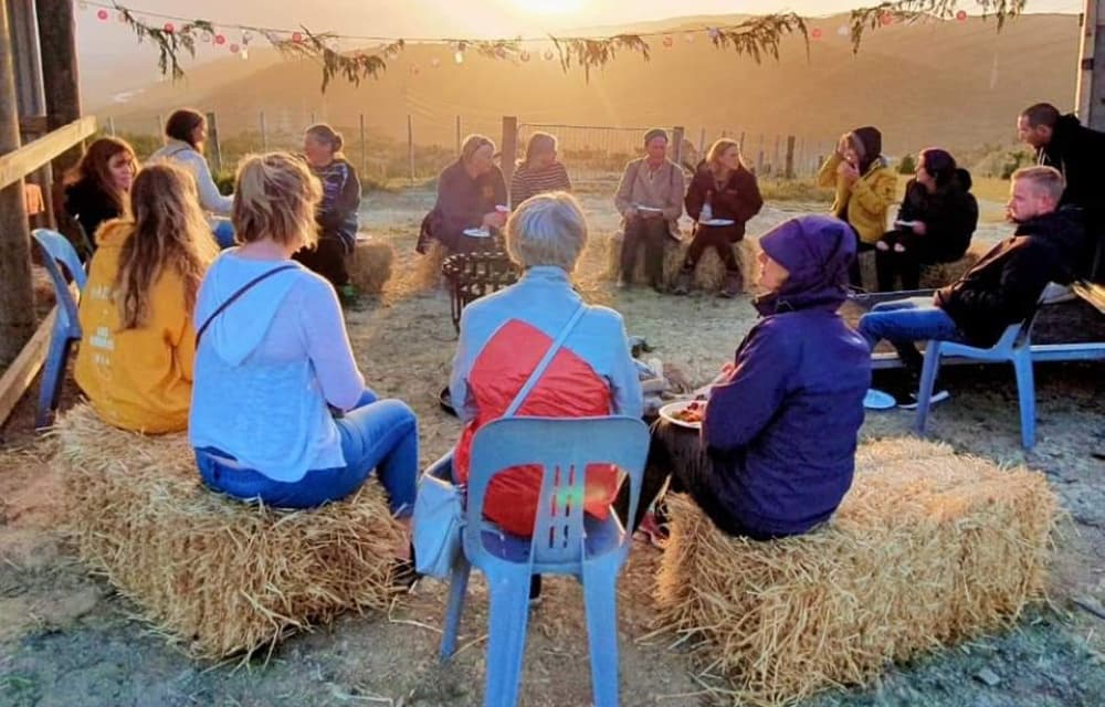Group of people in warm clothes gathered in a circle sitting on hay bales and plastic outdoor chairs with the sun setting in the background