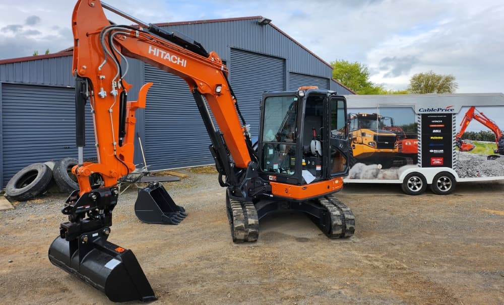Brand new small orange Hitachi digger parked up in front of a large blue garage building with a spare bucket to the side and heavy machinery in the background