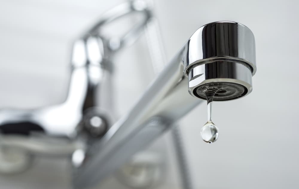 Close up of a shiny sink faucet with a big droplet of water coming out and a blurred background