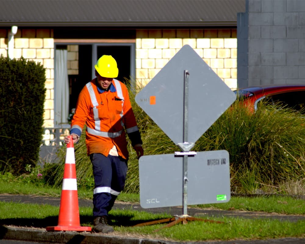Traffic management worker in orange high viz reflective gear setting up a cone next to a roadworks sign on the side of the road