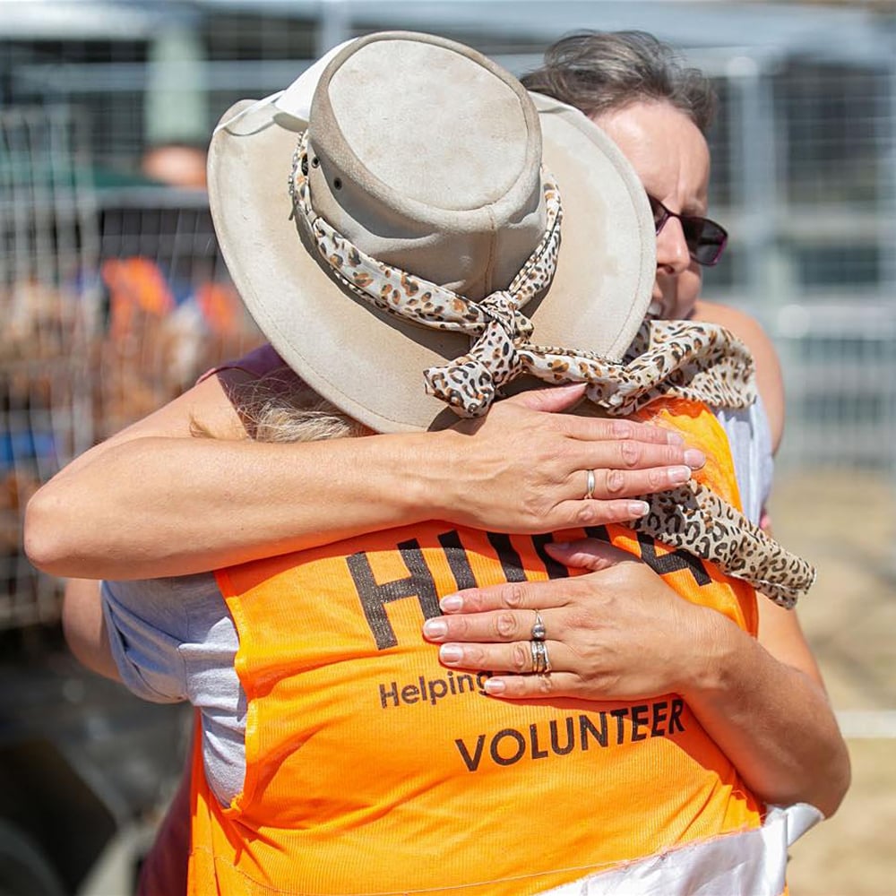 Huha volunteer in an orange high-viz vest and a tan cowboy hat giving a woman a hug