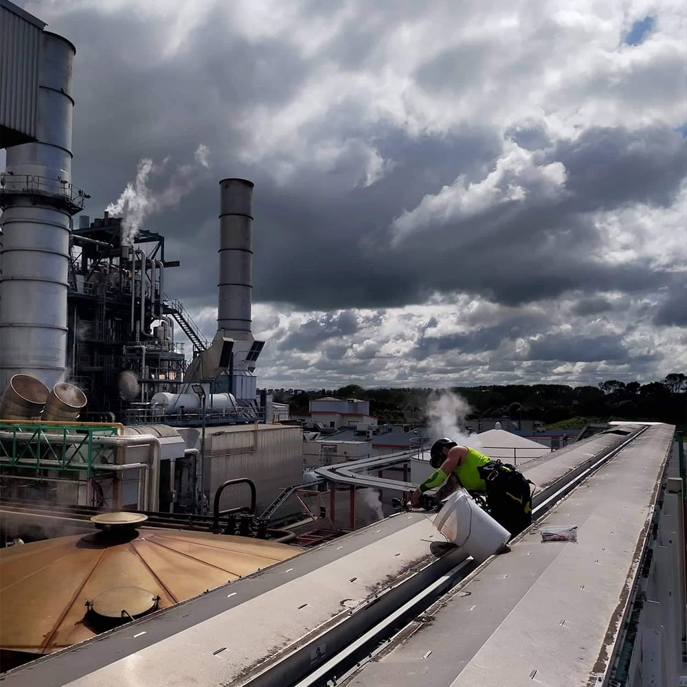 Maintenance guy on top of a factory building wall making repairs with a huge industrial factory in the background