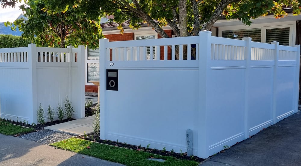 Flawless tall white fence giving privacy to the front yard of a small brick house with a concrete tile walkway and a couple medium trees