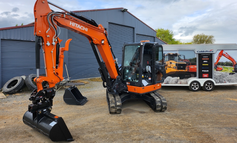 Brand new small orange Hitachi digger parked up in front of a large blue garage building with a spare bucket to the side and heavy machinery in the background