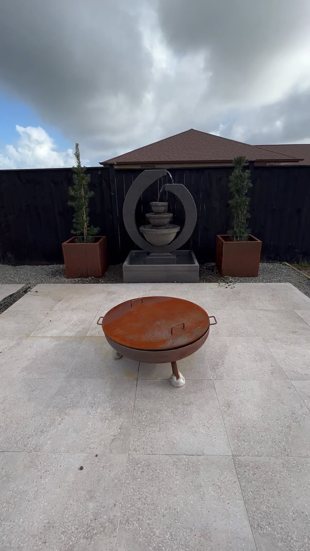 Outdoor firepit on a stone tile floor with two square flower pots and an articulate grey water fountain structure