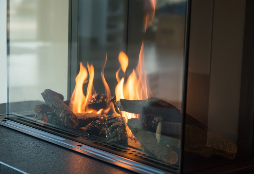 Large orange flames burning sizable pieces of wood behind the glass of a fireplace
