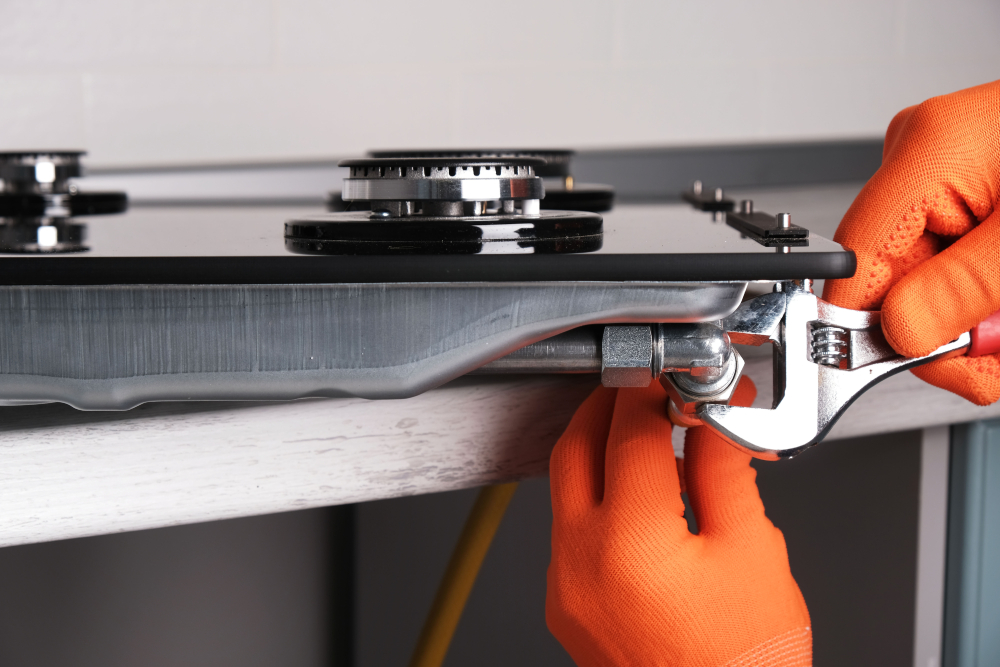Gasfitter with bright orange gloves using a small wrench to tighten a pipe under a gas stove