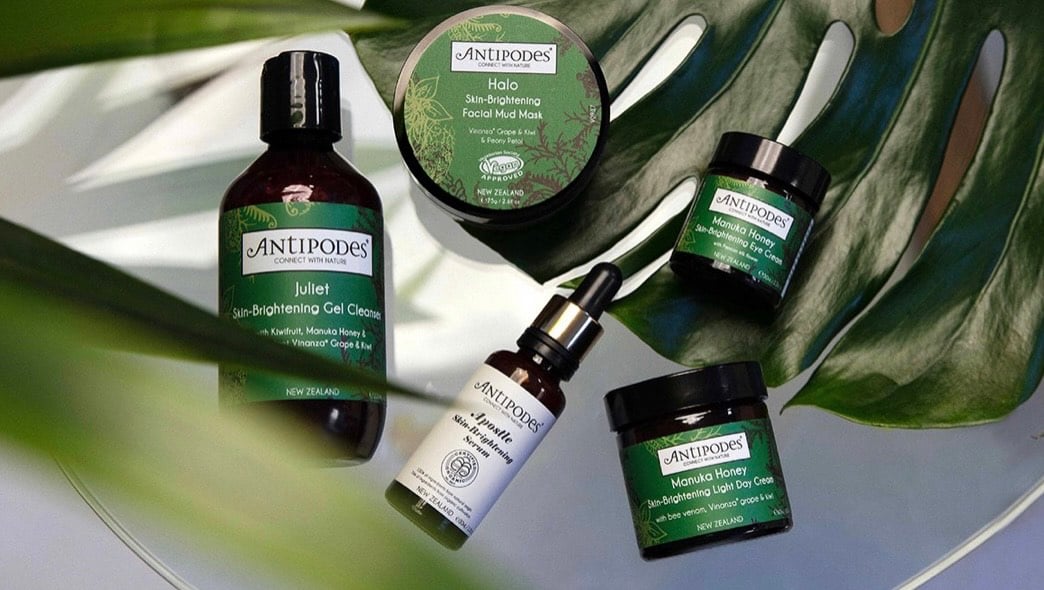 Range of different Antipodes skincare products with green labels on display on top of a monstera plant 