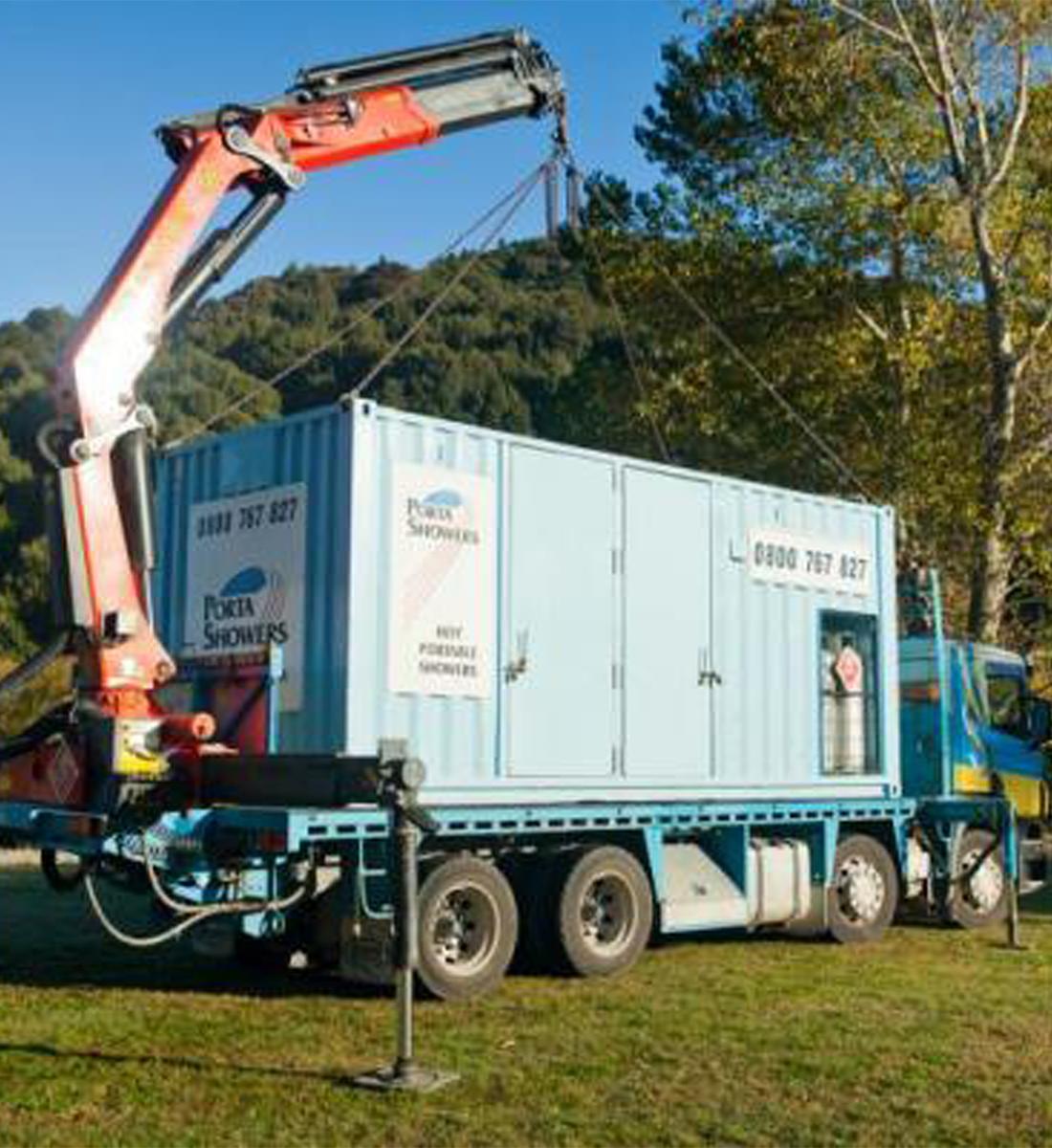 Truck crane lifting a Porta Showers container unit off the truck to set it down in a field of grass surrounded by tree-covered hills