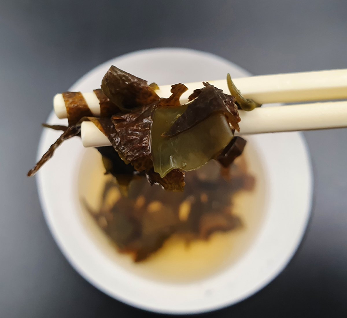 Chopsticks holding up small pieces of wakame seaweed above a white bowl of seaweed in water