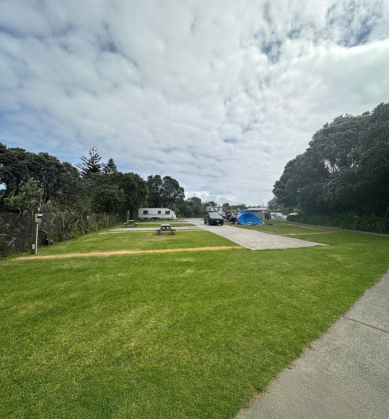 View of some empty powered camping sites at the Pinewoods Holiday Park. Flat, green grass and a large caravan in the background