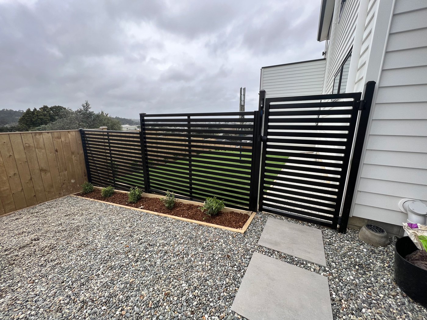 Modern black fence and gate that divide the yard from a pebble area with white stone tiles to walk on