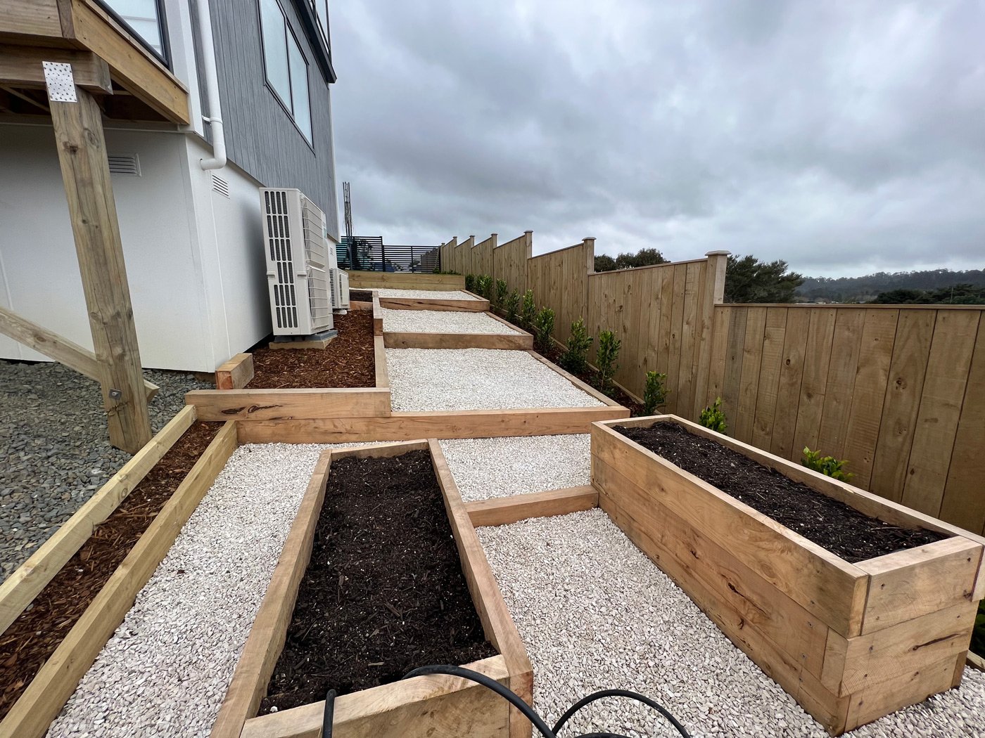 Large outside area going down the side of a newly built home with a couple vegetable beds and a row of freshly planted trees along the fence line