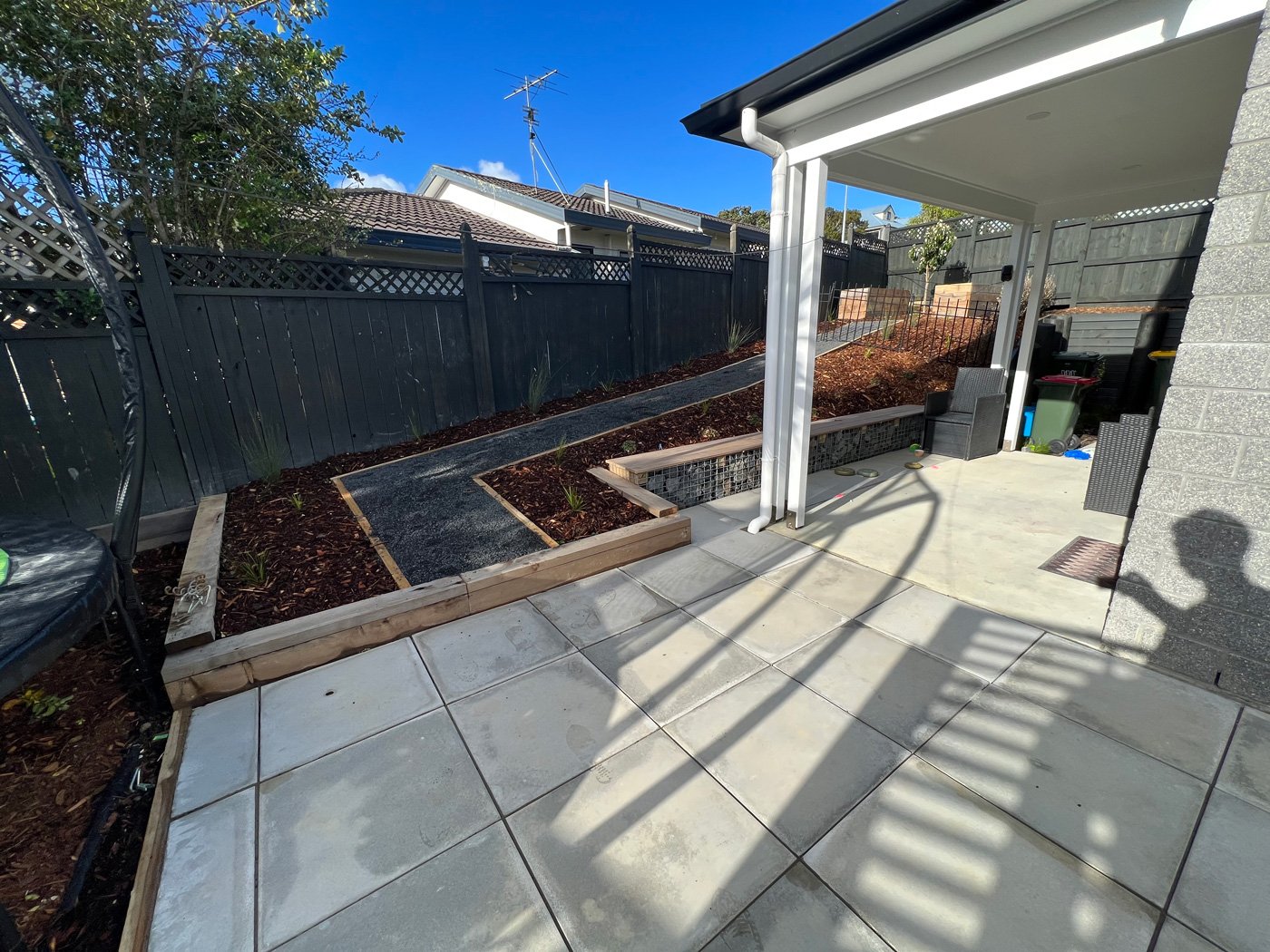 Concrete tiled backyard area with a gravel path outlined with wood and supported by a stone and wood retaining wall