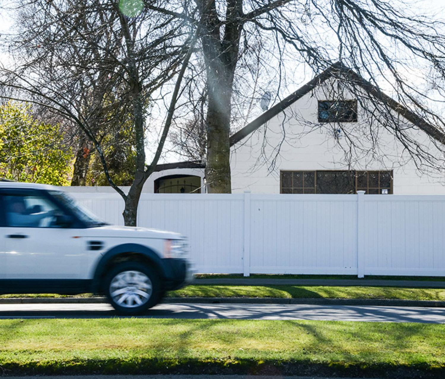 Moving vehicle in front of a tall white fence that offers privacy to a house with large windows