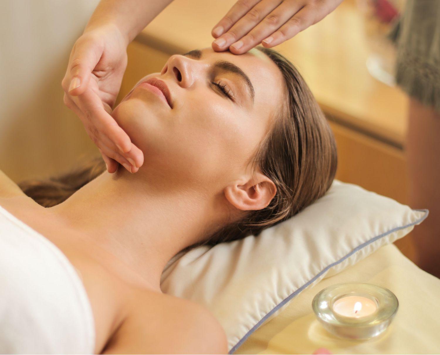 Woman wrapped in a towel, laying down on a comfy pillow with her eyes closed as someone massages her face