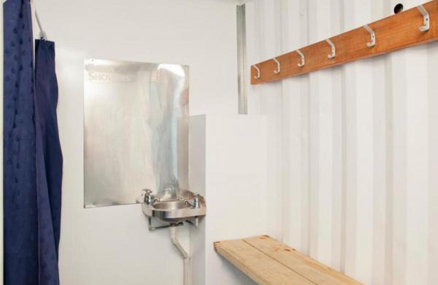 Porta Showers container unit interior with blue shower curtains, many hooks for hanging clothes or bags, and a small metal sink with a mirror