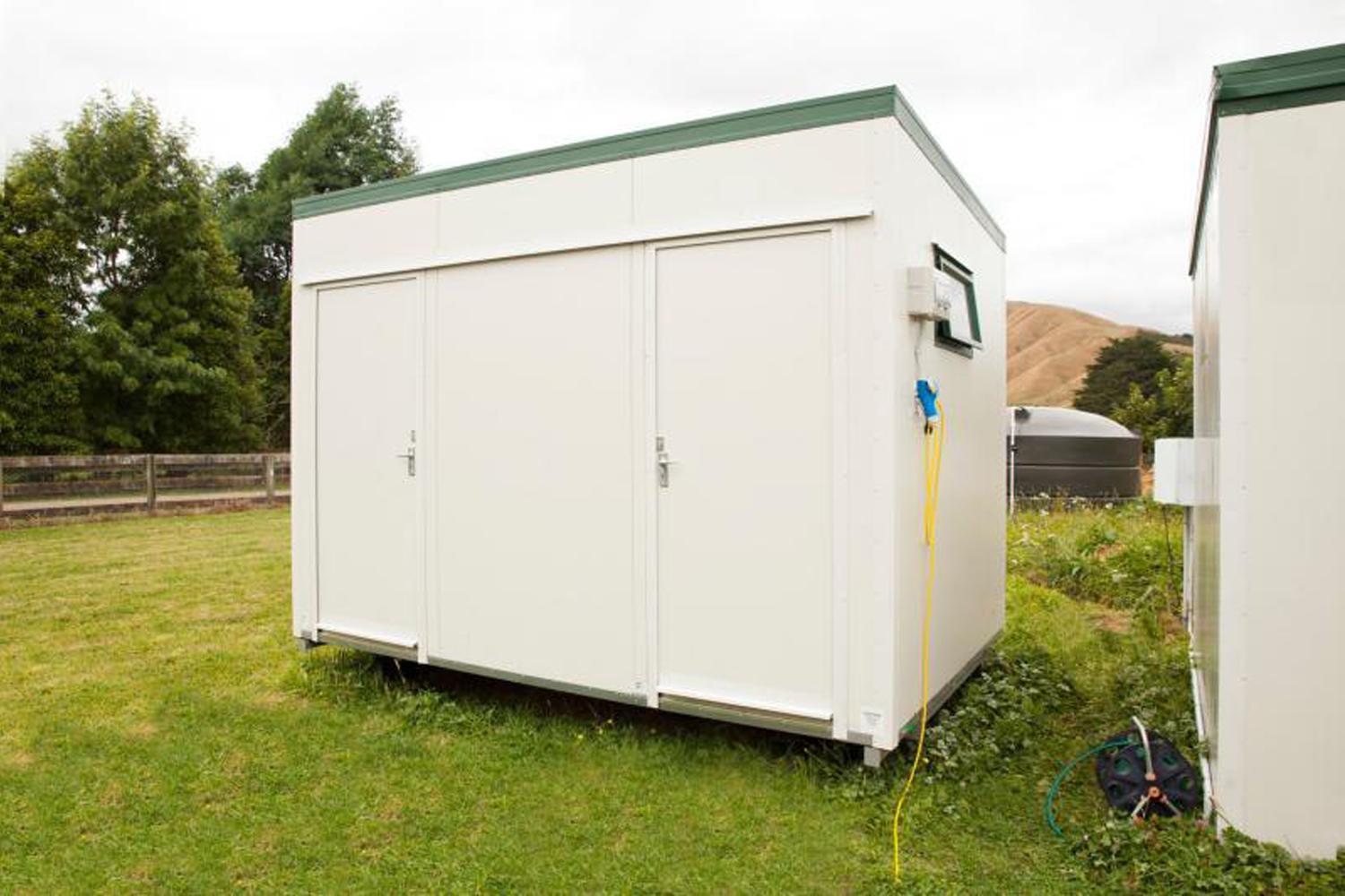 Big white utility unit shower block sat in a field with nature, a fence, and a water tank in the background