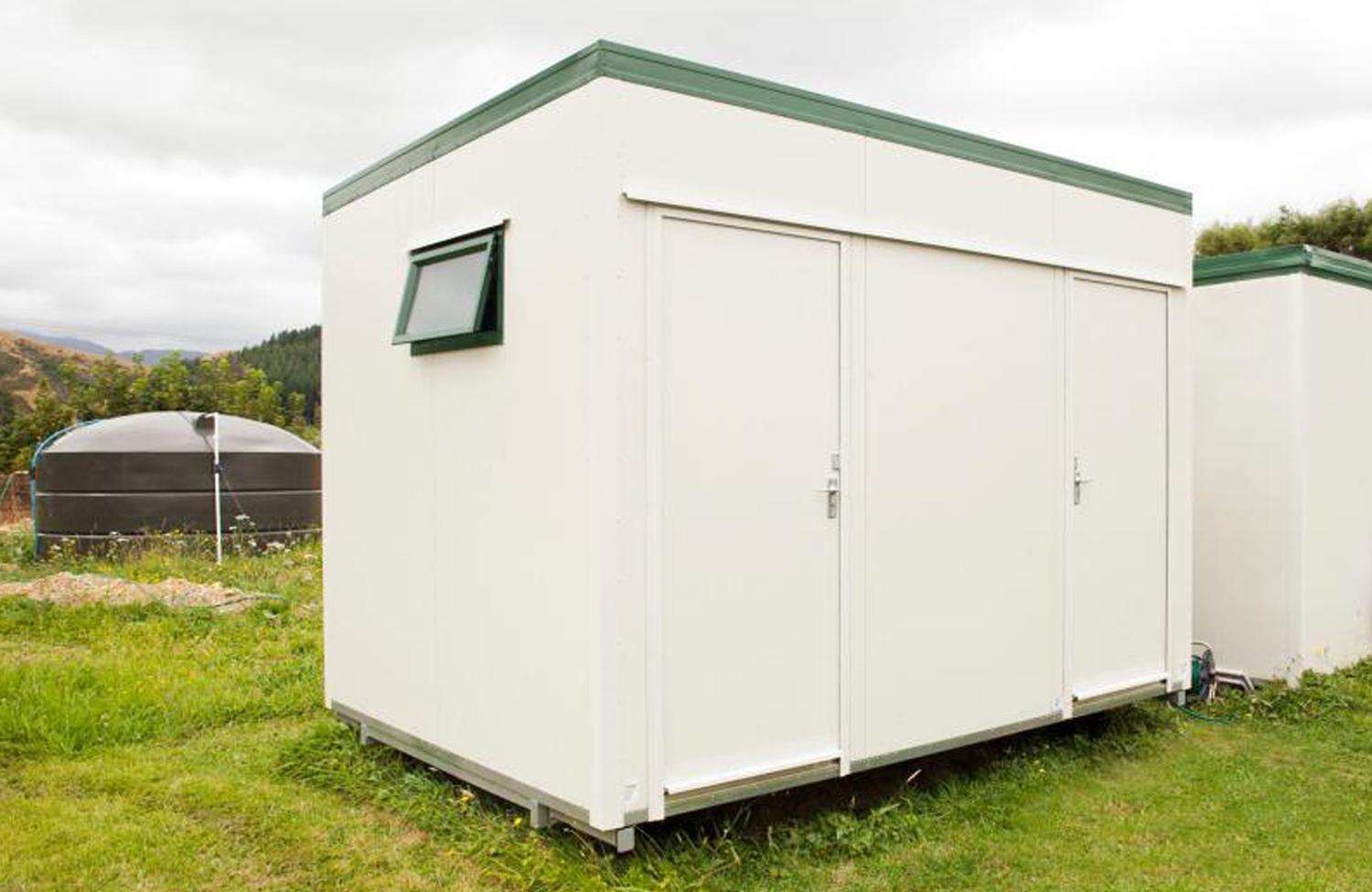 Big white utility unit shower block sat in a field with nature and a water tank in the background