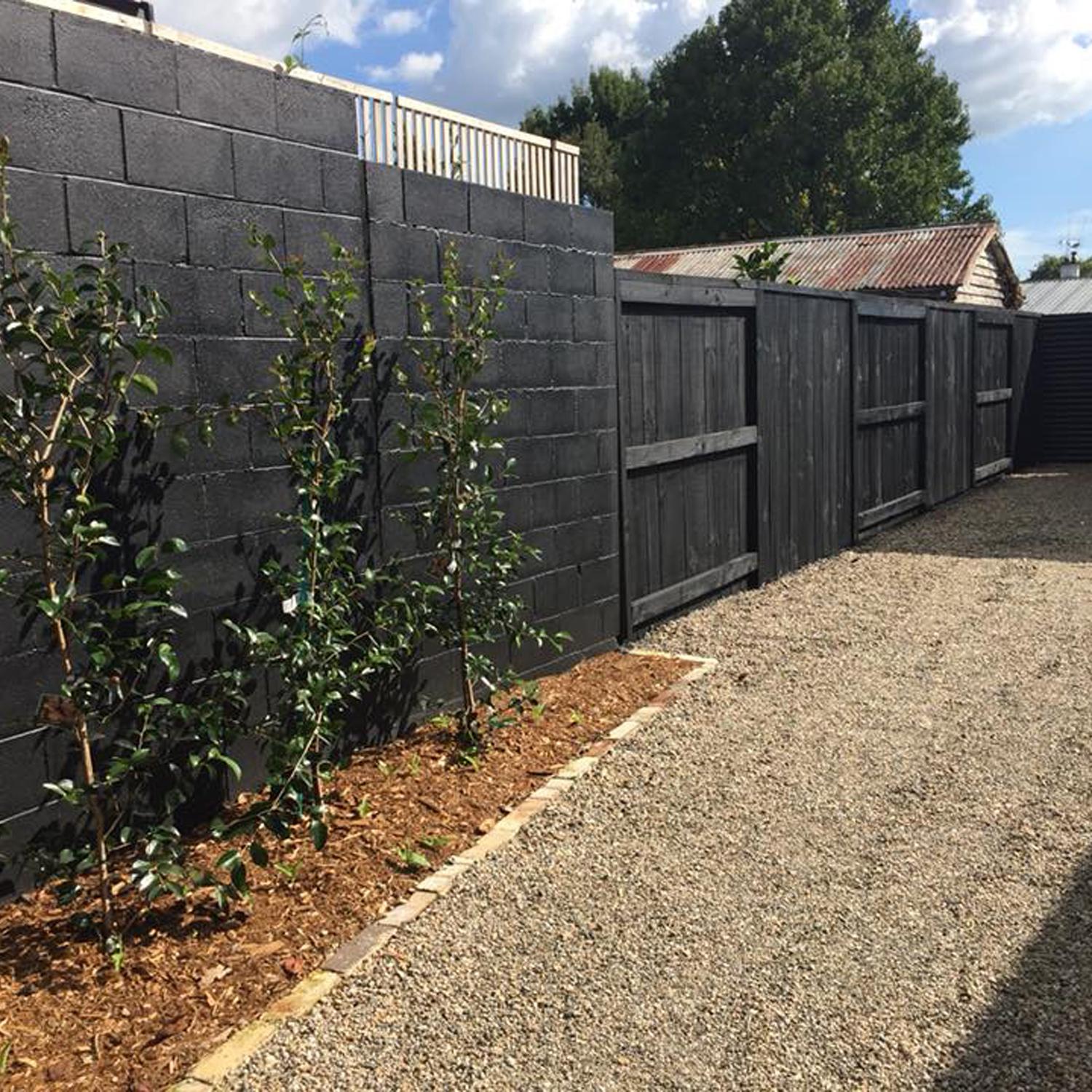 Black painted brick wall and wooden fence with tree saplings in a little mulch garden next to a gravel driveway