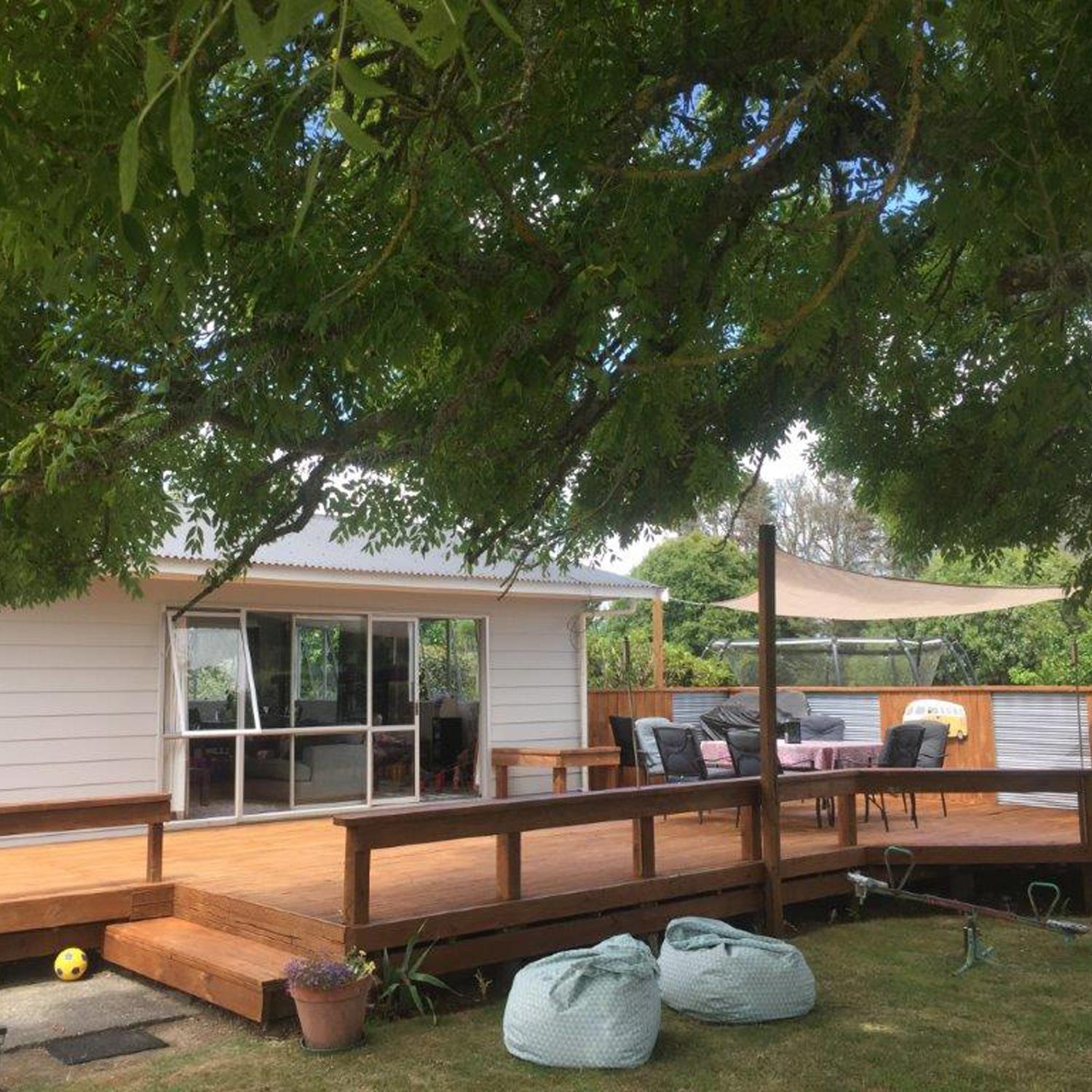 Spacious wooden deck stained beautifully with a canopied seating area and a couple bean bags on the grass under a large tree 