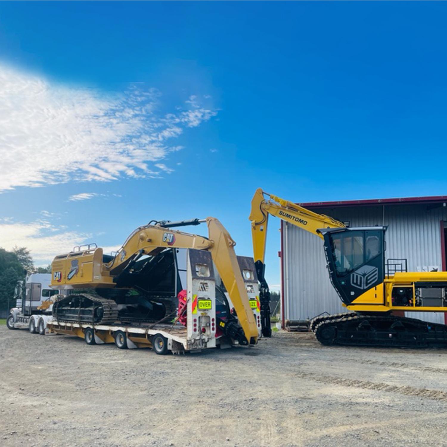 A long white truck transporting a large yellow digger with another large yellow digger parked up next to a rural factory building