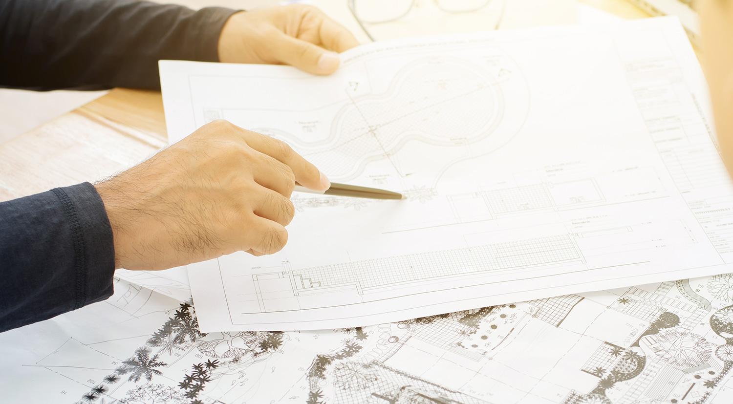 Close up view of a landscape architects hands holding a pen and pointing to a specific piece on a blue print design paper - links to the Why Get Independent Advice page