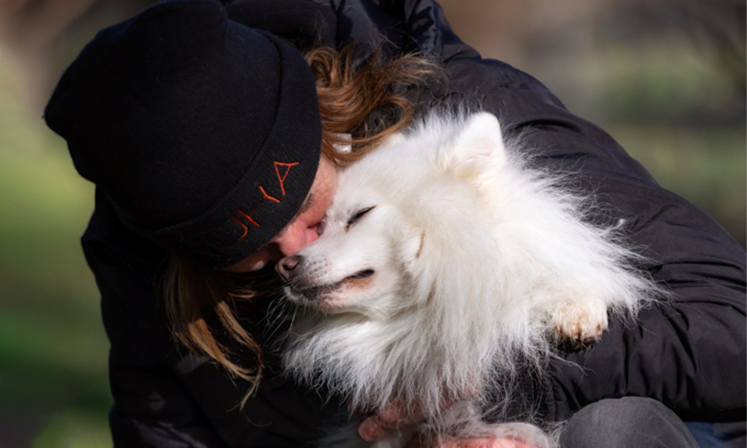 Lady in a warm jacket and Huha beanie giving a fluffy white dog a big hug in the middle of a field
