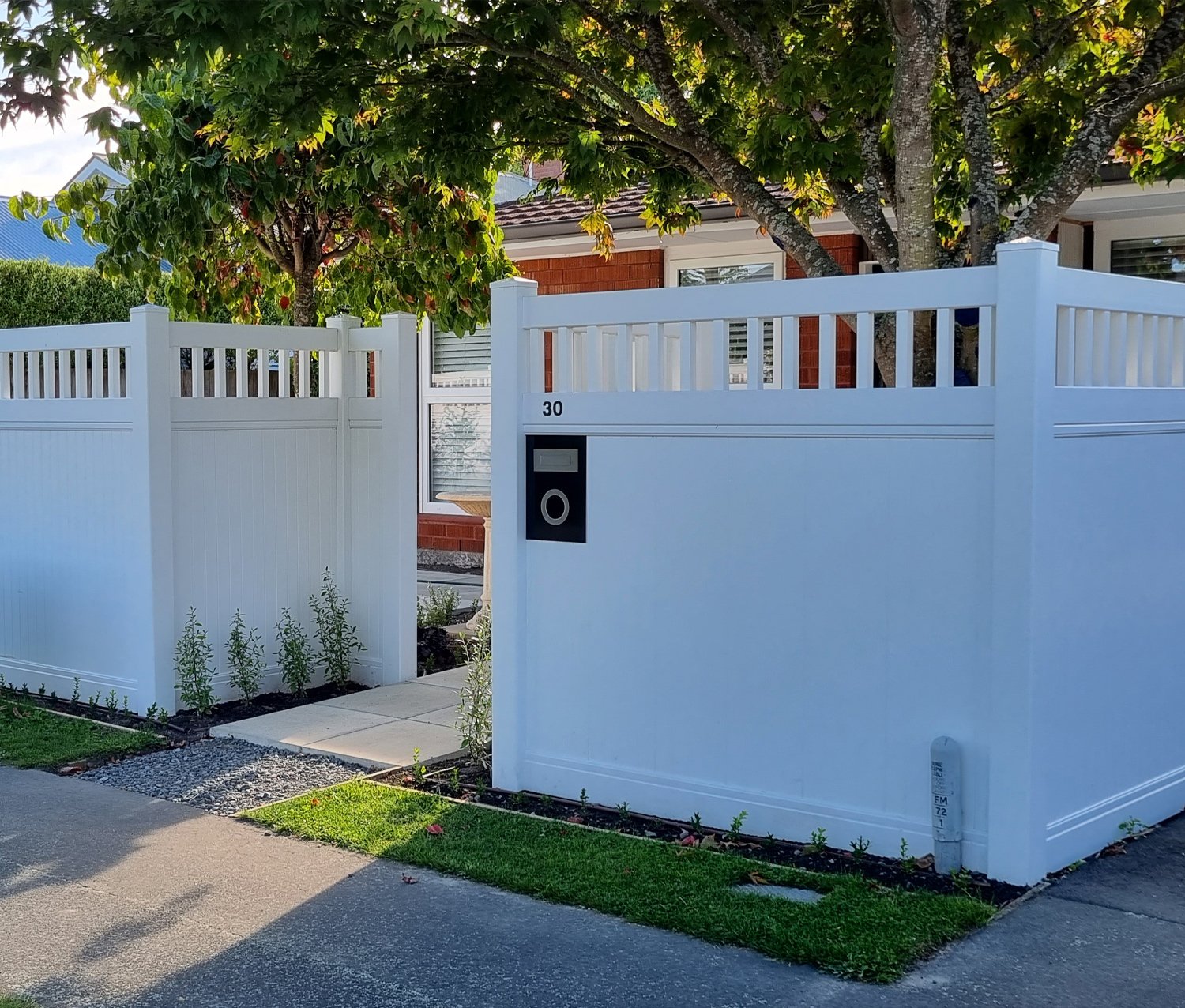 Flawless tall white fence giving privacy to the front yard of a small brick house with a concrete tile walkway and a couple medium trees