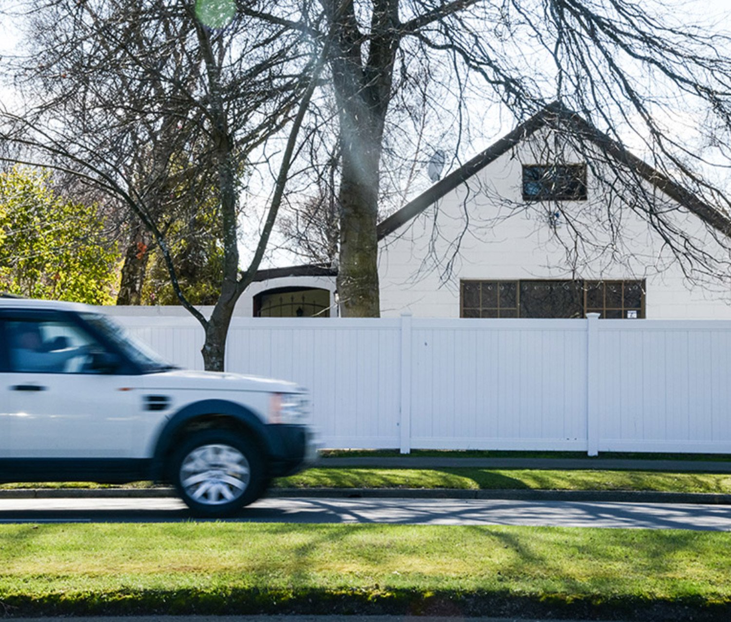 Moving vehicle in front of a tall white fence that offers privacy to a house with large windows