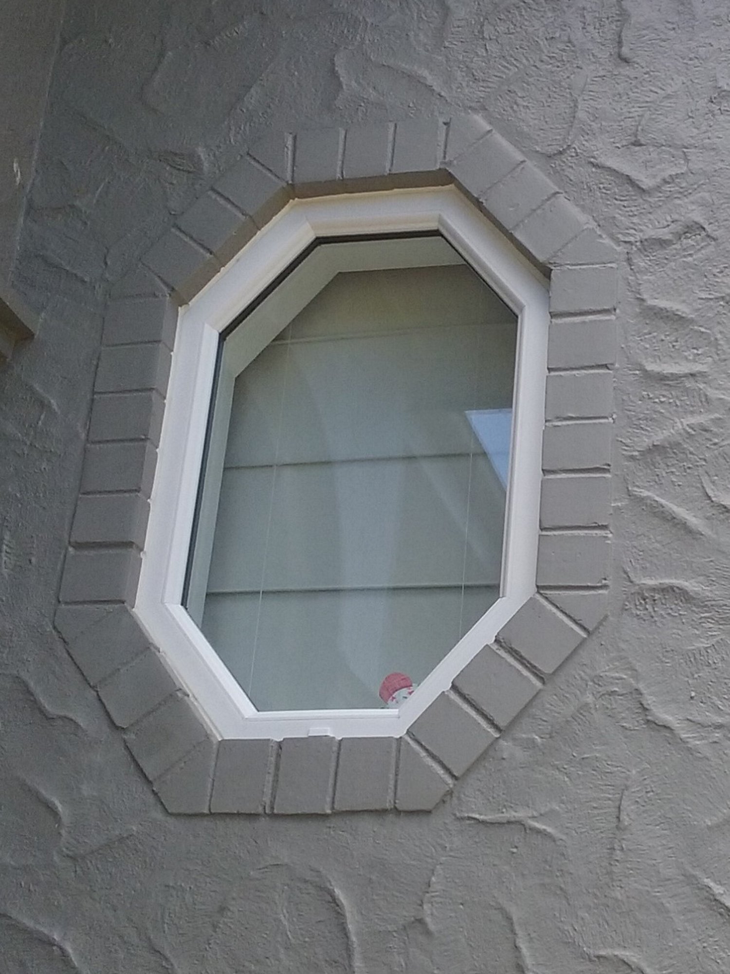 Small octagonal window with a white upvc window frame on a grey paved stone wall 