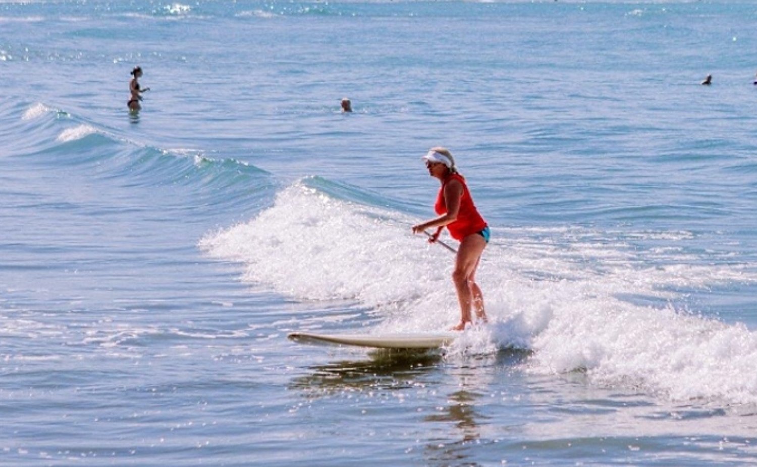 Picture of a Lady in a pink bathing suit surfing a gentle wave on her stand-up paddle board while holding an ore. She is at the local Red beach