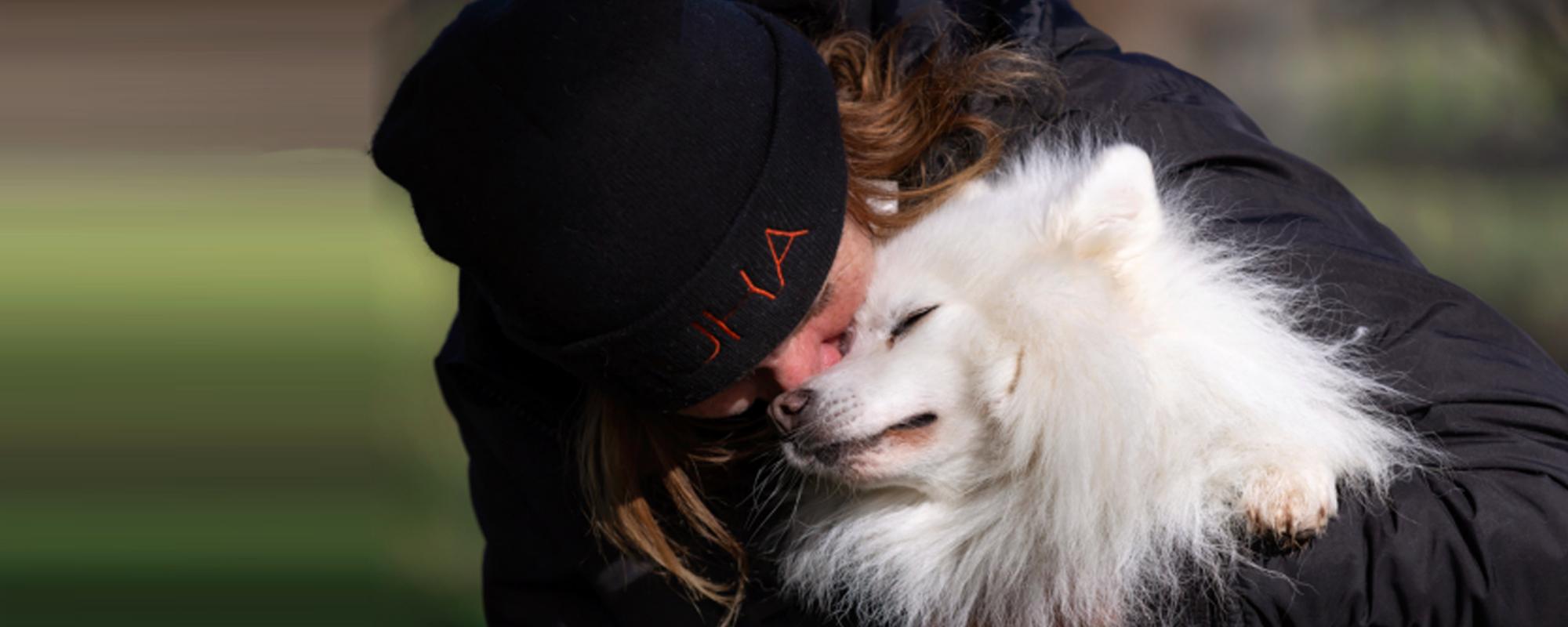 Lady in a warm jacket and Huha beanie giving a fluffy white dog a big hug in the middle of a field