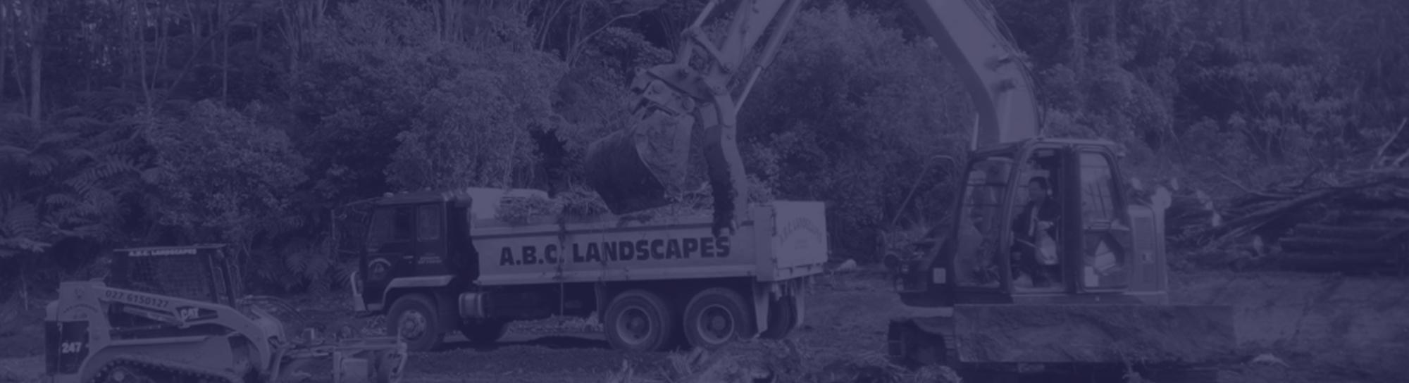 Large orange digger moving timber debris from the site into an ABC Landscapes truck on a partially cleared site