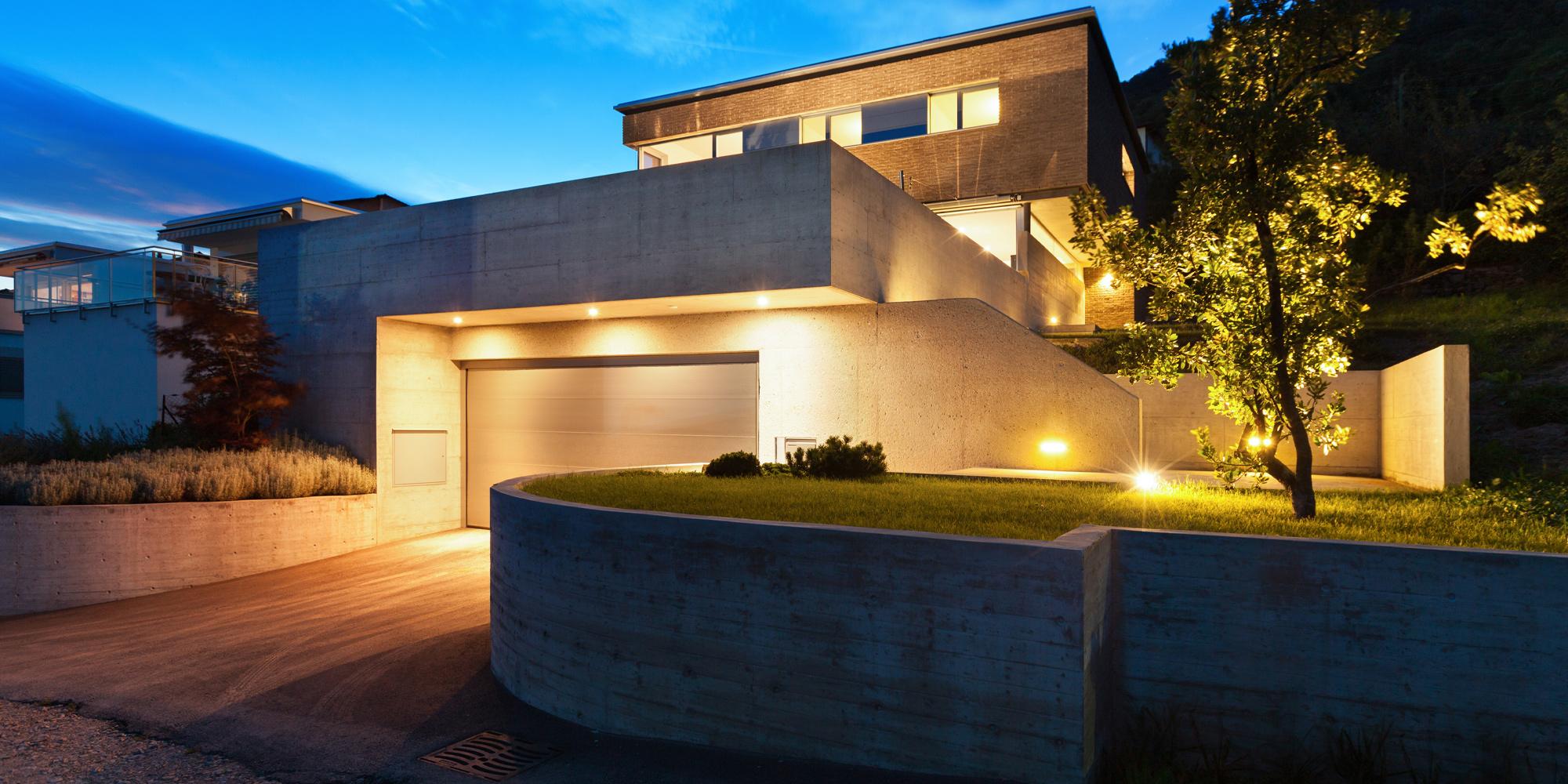 Beautifully lit up residential property with amazing concrete architecture, grey double garage door, and a raised front garden with a concrete retaining wall