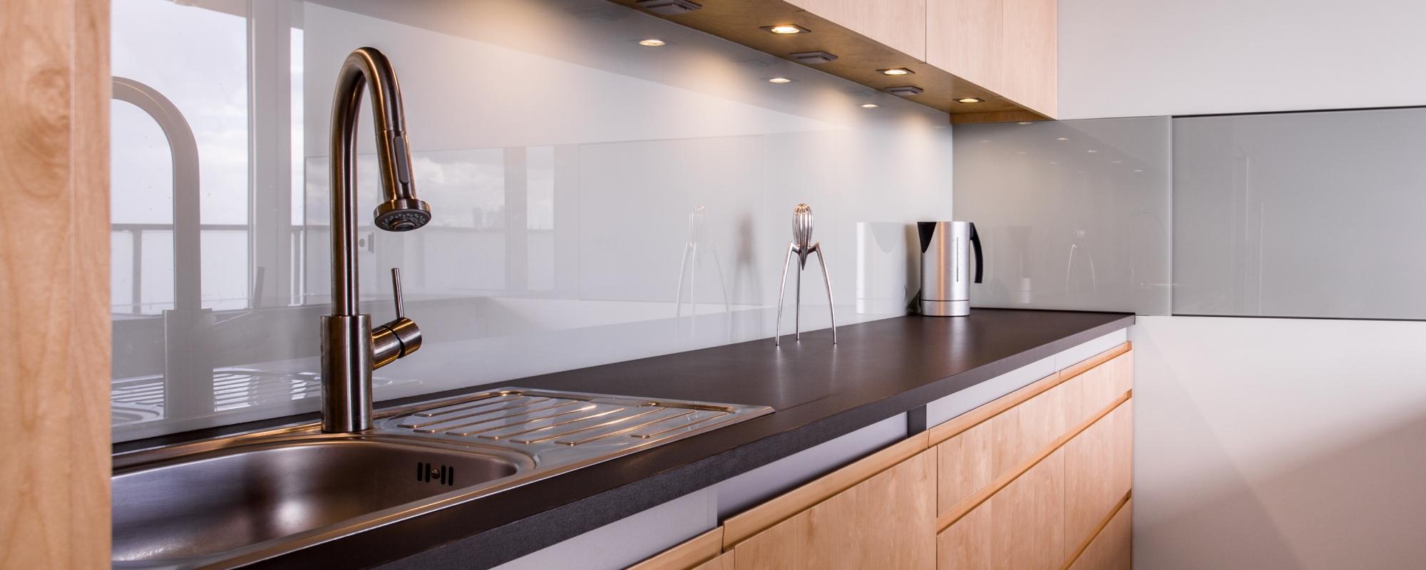 Kitchen with wooden cabinetry, a matte-like black bench and a glossy white splashback reflecting in the light