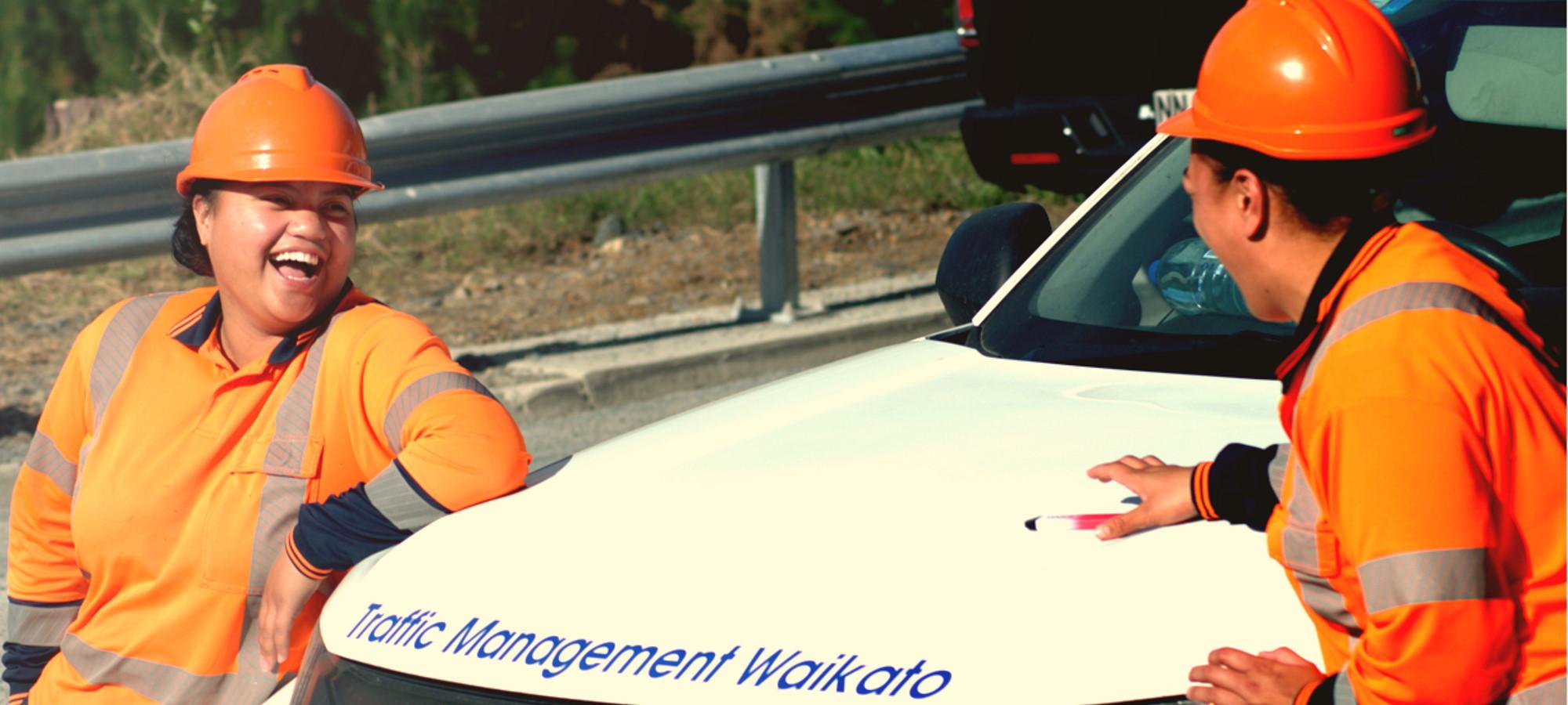 Two traffic management waikato team members leaned up against their work vehicle having a laugh while dressed in their high viz orange work gear