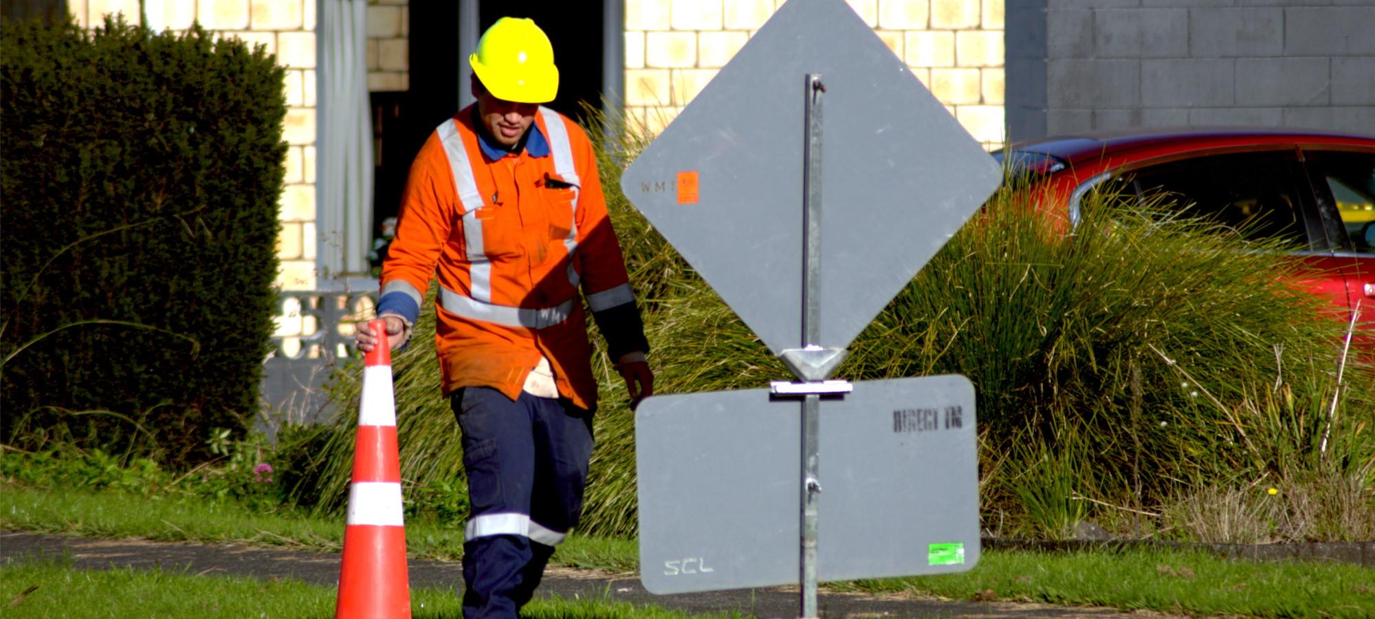 Traffic management worker in orange high viz reflective gear setting up a cone next to a roadworks sign on the side of the road
