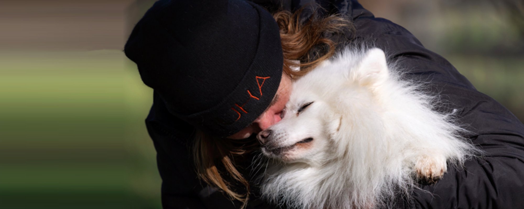 Lady in a warm jacket and Huha beanie giving a fluffy white dog a big hug in the middle of a field