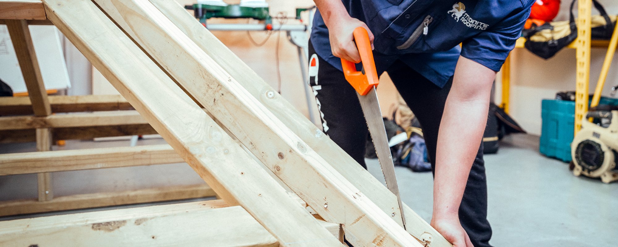 Midnite roofing apprentice using an orange-handled saw to cut excess wood off a pre-made wood roof frame