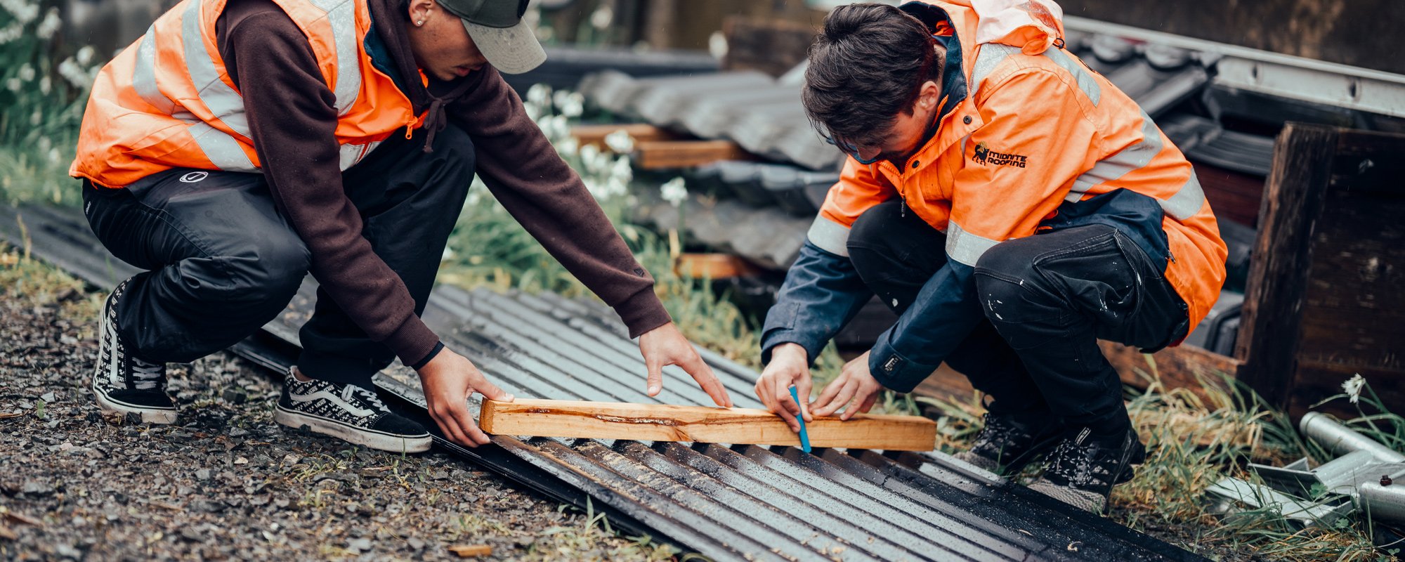 Two roofers from Midnite Roofing using a piece of wood as a ruler to draw a straight line on a piece of black tin roofing