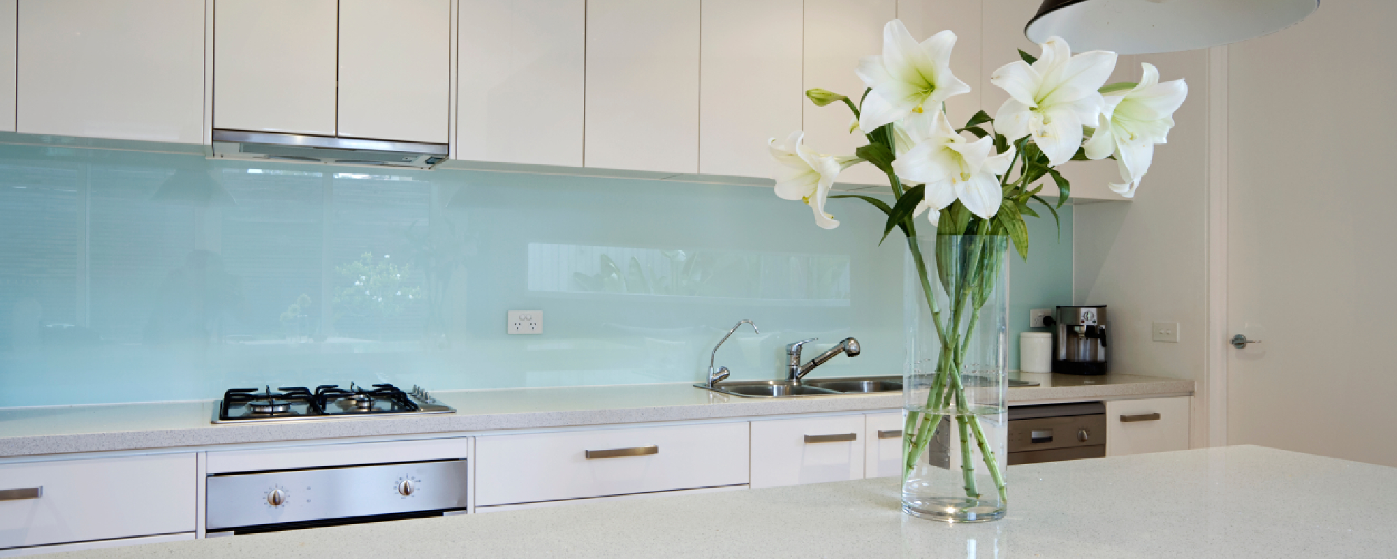 White kitchen cabinetry and benchtops with a reflective baby blue splashback and a tall vase with white lilys on the kitchen island