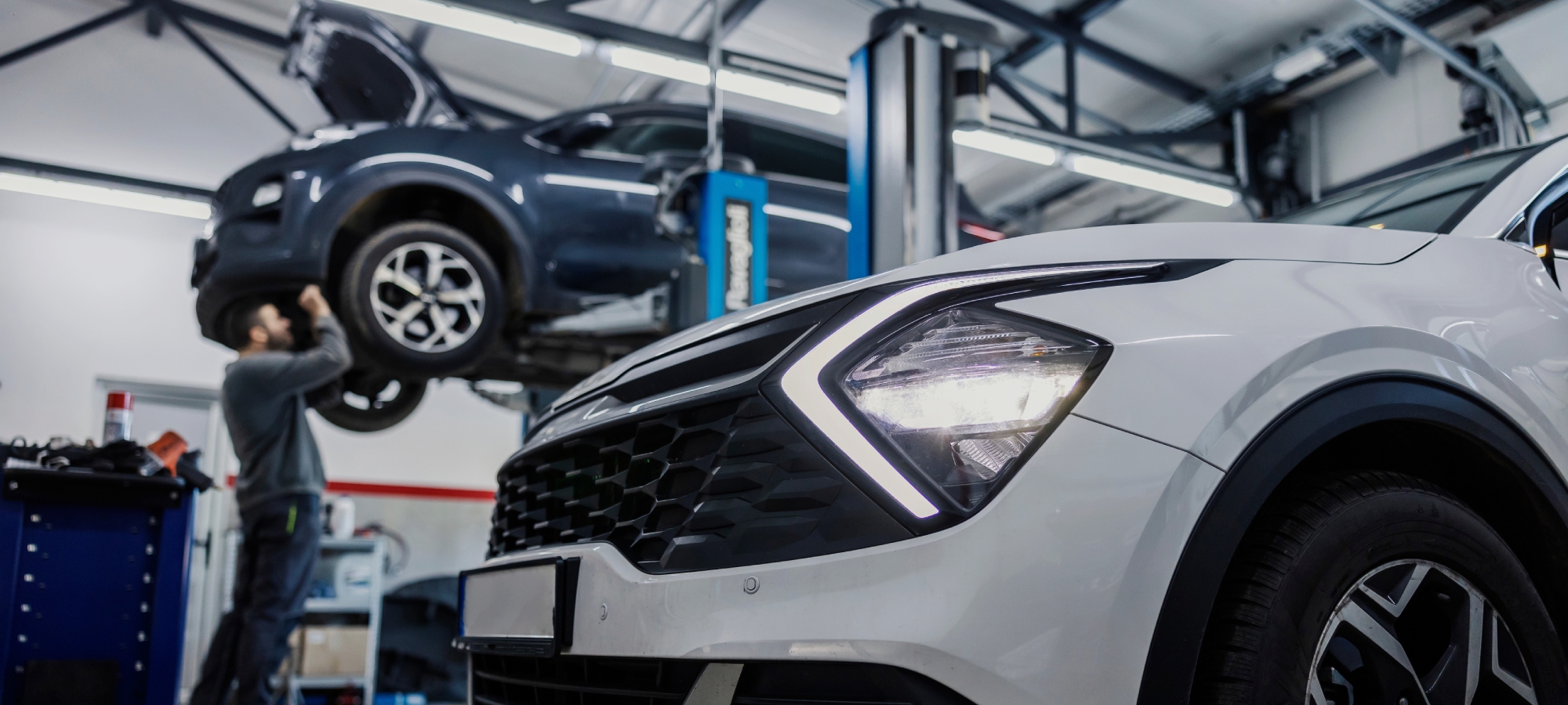 Mechanic's workshop with two cars, one parked up and one elevated with it's hood open and a mechanic working on the front of it
