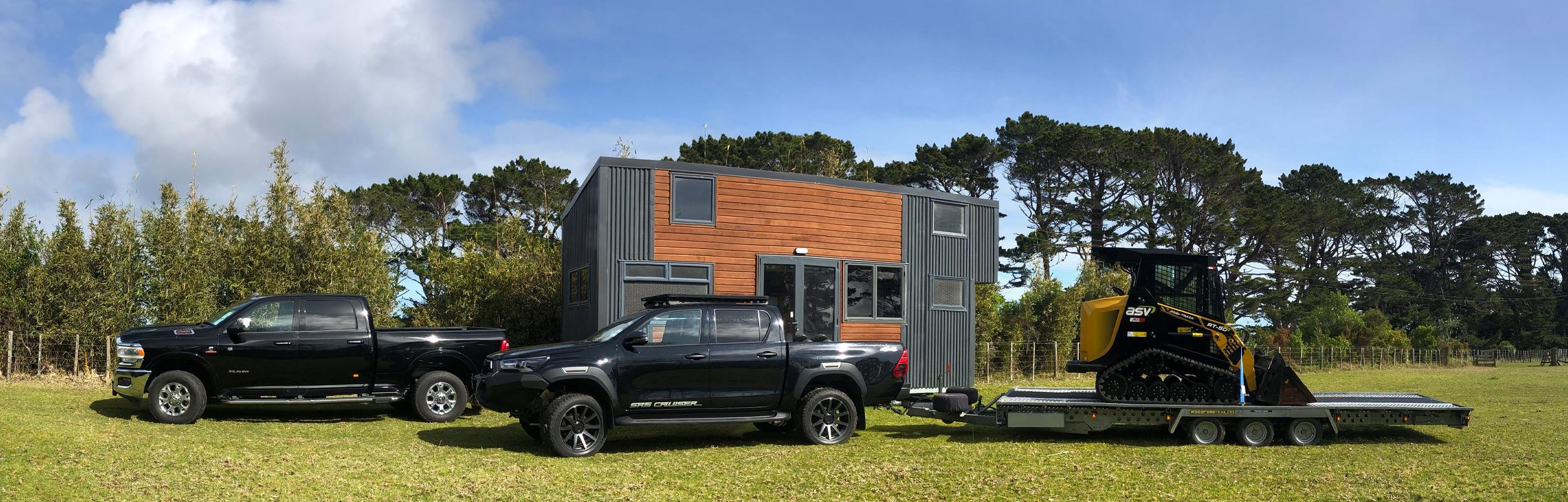 Two of NZ Tiny Home Movers' trucks towing a tiny home and a piece of heavy machinery for preparing sites