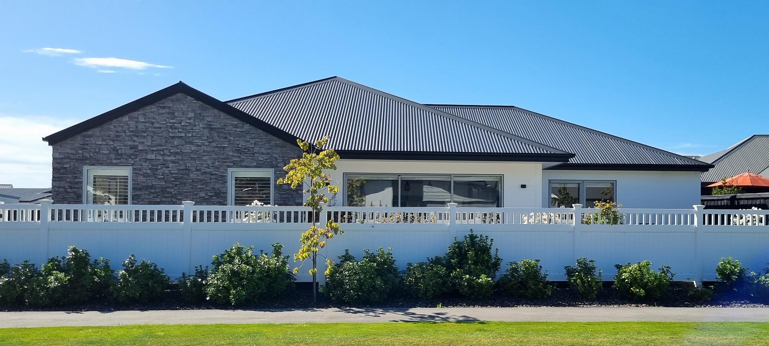 New white fencing with tree and bush saplings in front of a modern house with white painted exterior and a modern rock feature wall