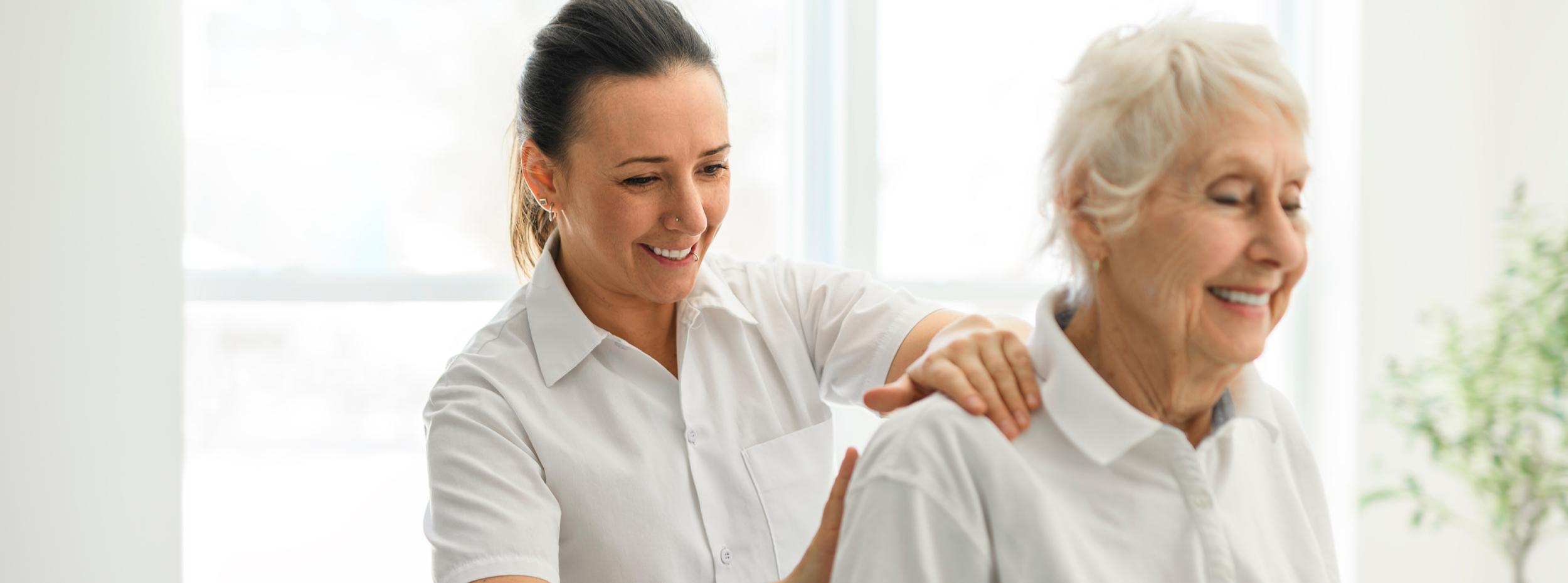 Female Osteopath treating an eldery female patient's shoulder. Standing behind smiling patient - Osteopaths North Shore Auckland