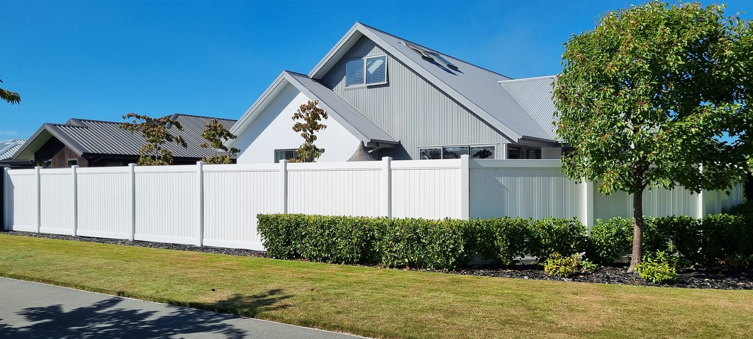 Brand new flawless tall white PVC fencing with trimmed hedges and surrounding trees in front of a two story white and grey house